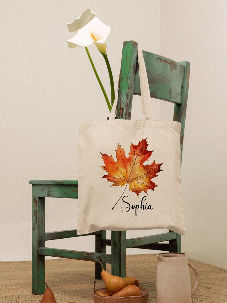 May include: A white canvas tote bag featuring a watercolour illustration of an orange and yellow maple leaf. The bag is inscribed with the name "Sophia" in cursive below the leaf. The bag is hanging on a green wooden chair.