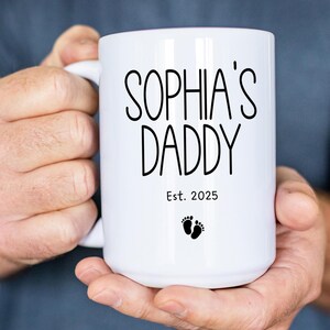 Peut inclure: Une tasse en c&eacute;ramique blanche avec du texte noir qui dit "Sophia's Daddy Est. 2025" et un petit graphique de pied.
