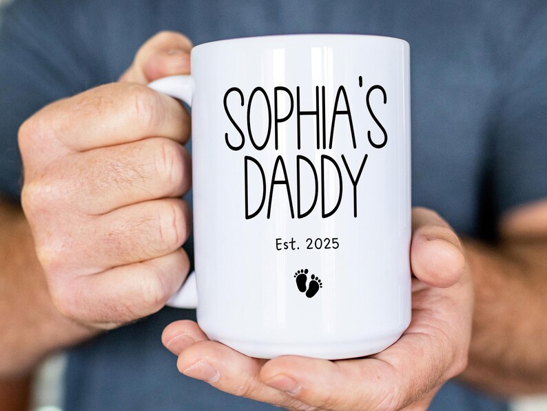 Peut inclure: Une tasse en c&eacute;ramique blanche avec du texte noir qui dit "Sophia's Daddy Est. 2025" et un petit graphique de pied.