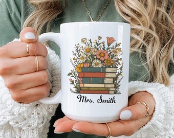 May include: White ceramic mug featuring a floral design above a stack of books. The flowers include daisies, pink and yellow blooms. The mug has the name "Mrs. Smith" printed on it.