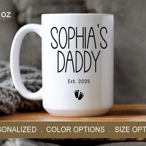 Peut inclure: Une tasse en c&eacute;ramique blanche avec du texte noir qui dit "Sophia's Daddy Est. 2025" et un petit graphique de pied. La tasse est de 15 onces.