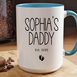 Peut inclure: Une tasse en c&eacute;ramique blanche avec un bord et une poign&eacute;e bleus. La tasse porte une inscription noire "Sophia's Daddy Est. 2025" et un petit symbole de pied.