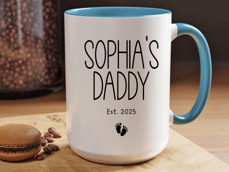 Peut inclure: Une tasse en c&eacute;ramique blanche avec un bord et une poign&eacute;e bleus. La tasse porte une inscription noire "Sophia's Daddy Est. 2025" et un petit symbole de pied.