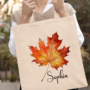 May include: A beige tote bag featuring a watercolour painting of an orange and yellow maple leaf. The name "Sophia" is written in black cursive below the leaf.