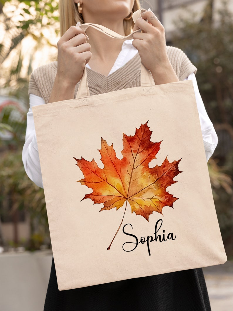 May include: A beige tote bag featuring a watercolour painting of an orange and yellow maple leaf. The name "Sophia" is written in black cursive below the leaf.