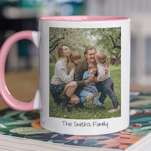 Puede incluir: Una taza de cer&aacute;mica blanca con un borde y un asa de color rosa. La taza presenta una foto de una familia de cuatro personas en un entorno de jard&iacute;n. El texto "The Smith's Family" est&aacute; impreso en la parte inferior de la taza.