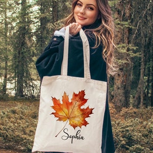 May include: A white canvas tote bag featuring a watercolour illustration of an orange and yellow maple leaf. The bag is personalised with the name "Sophia" written in cursive below the leaf.