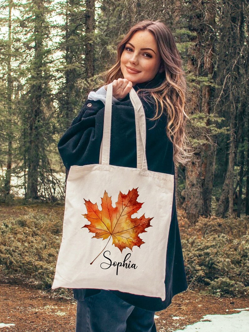 May include: A white canvas tote bag featuring a watercolour illustration of an orange and yellow maple leaf. The bag is personalised with the name "Sophia" written in cursive below the leaf.