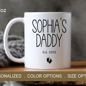 Peut inclure: Une tasse en c&eacute;ramique blanche avec du texte noir qui dit "Sophia's Daddy Est. 2025" et deux petites empreintes de pas noires. La tasse est de 11 onces.
