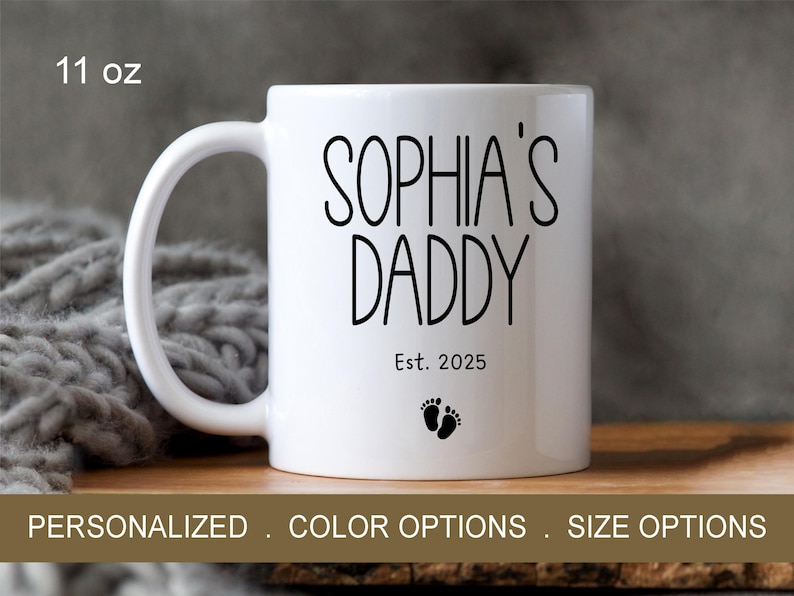 Peut inclure: Une tasse en c&eacute;ramique blanche avec du texte noir qui dit "Sophia's Daddy Est. 2025" et deux petites empreintes de pas noires. La tasse est de 11 onces.