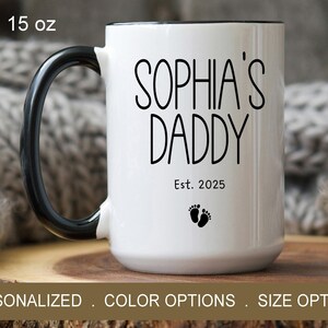 Peut inclure: Une tasse en c&eacute;ramique blanche avec une anse et un bord noirs. La tasse porte l'inscription "Sophia's Daddy Est. 2025" avec deux petites empreintes de pas noires en dessous du texte. La tasse a une contenance de 15 onces.