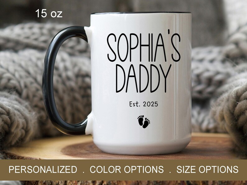 Peut inclure: Une tasse en c&eacute;ramique blanche avec une anse et un bord noirs. La tasse porte l'inscription "Sophia's Daddy Est. 2025" avec deux petites empreintes de pas noires en dessous du texte. La tasse a une contenance de 15 onces.