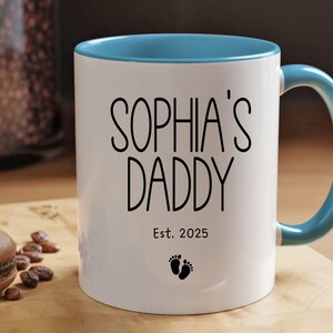 Peut inclure: Une tasse en c&eacute;ramique blanche avec un bord et une poign&eacute;e bleu clair. La tasse porte une inscription noire "Sophia's Daddy Est. 2025" et un petit graphique de pied.