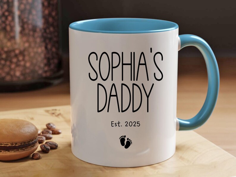 Peut inclure: Une tasse en c&eacute;ramique blanche avec un bord et une poign&eacute;e bleu clair. La tasse porte une inscription noire "Sophia's Daddy Est. 2025" et un petit graphique de pied.