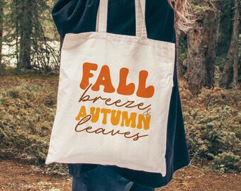 May include: A white tote bag with the text "FALL breeze & autumn leaves" in orange and yellow lettering. The bag is being held by a person in a forest setting.