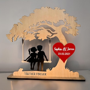 May include: A wooden decorative piece featuring a tree with a swing holding two silhouettes of people kissing. A red heart with the names "Sophia & James" and the date "23.02.2021" is attached to the tree. The base reads "TOGETHER FOREVER."