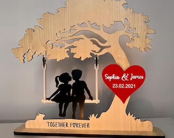 Puede incluir: Adorno de madera que representa un árbol con un columpio con dos siluetas besándose. Un corazón rojo con los nombres "Sophia & James" y la fecha "23.02.2021" está adjunto. La base dice "TOGETHER FOREVER."