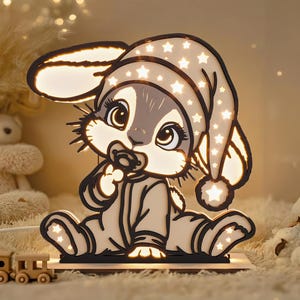 Puede incluir: Una luz de noche decorativa con forma de conejito bebé que lleva un gorro de dormir con recortes de estrellas. El conejito es gris y blanco, con un chupete y está sentado sobre una base de madera. La luz está rodeada de otros juguetes.