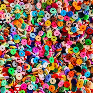May include: A close-up of a collection of colourful, flat, circular beads with holes. The beads are in a variety of shades, including pink, orange, yellow, green, blue, and white. The image presents a textured and visually appealing arrangement of the beads.