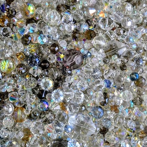 May include: A close-up shot of a collection of clear and iridescent crystal beads. The beads are of various sizes and shapes, with facets that reflect light, creating a sparkling effect. The colour palette includes clear, brown, and blue tones.