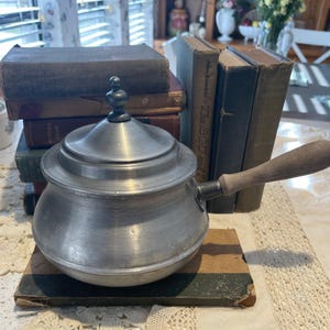 May include: A silver metal saucepan with a wooden handle and a lid. The saucepan is sitting on a stack of old books.