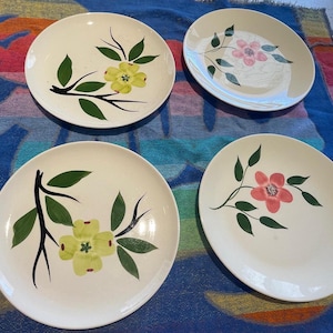 May include: Four white ceramic dinner plates with floral designs. Two plates feature pink flowers with green leaves, and two plates feature yellow flowers with green leaves.