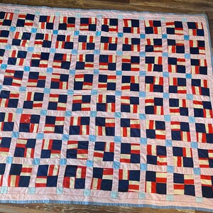 May include: A patchwork quilt with a grid pattern of squares in pink, blue, white, and red. The quilt is made of fabric scraps sewn together in a random pattern.