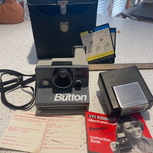 May include: A gray Polaroid Land Camera called "The Button" with a black case, a flash unit, and an instruction booklet. The instruction booklet is red and white and has the text "ITT Magicflash Electronic Flash for Polaroid Pronto! Land Camera Instruction Book".