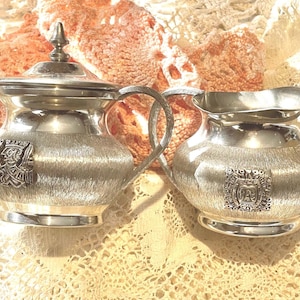 May include: A silver sugar and creamer set with ornate designs. The pieces have a brushed finish and feature a raised crest with a stylized figure.