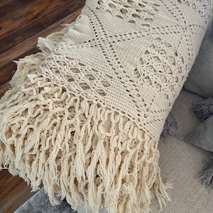May include: A cream-colored crocheted throw blanket with a fringe border. The blanket is made with a delicate openwork pattern and has a soft, textured feel.