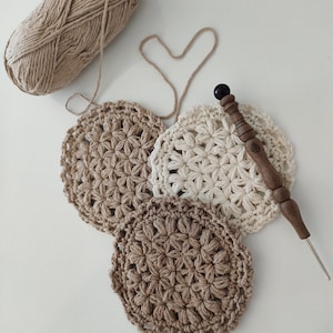 Könnte beinhalten: Drei gehäkelte Untersetzer in Beige und Weiß mit einer Häkelnadel aus Holz. Die Untersetzer sind mit einem sternförmigen Stich gearbeitet.