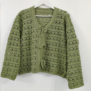 May include: An olive green crochet cardigan with a V-neck and long sleeves. The openwork design features a delicate pattern, and the front closure has three tie closures. The cardigan is hanging on a white hanger against a white background.
