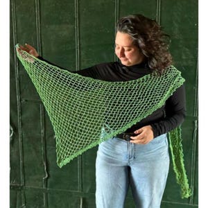 Könnte beinhalten: Ein grüner Häkel-Schal mit einem netzartigen Muster. Der Schal ist über die Schultern einer Person gelegt und wird mit einem schwarzen Langarmshirt und einer blauen Jeans getragen.