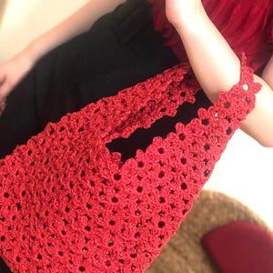 May include: A red crochet tote bag with a floral pattern. The bag is held by a person wearing a red top and black pants. The bag's handle is adorned with flower-shaped crochet details.