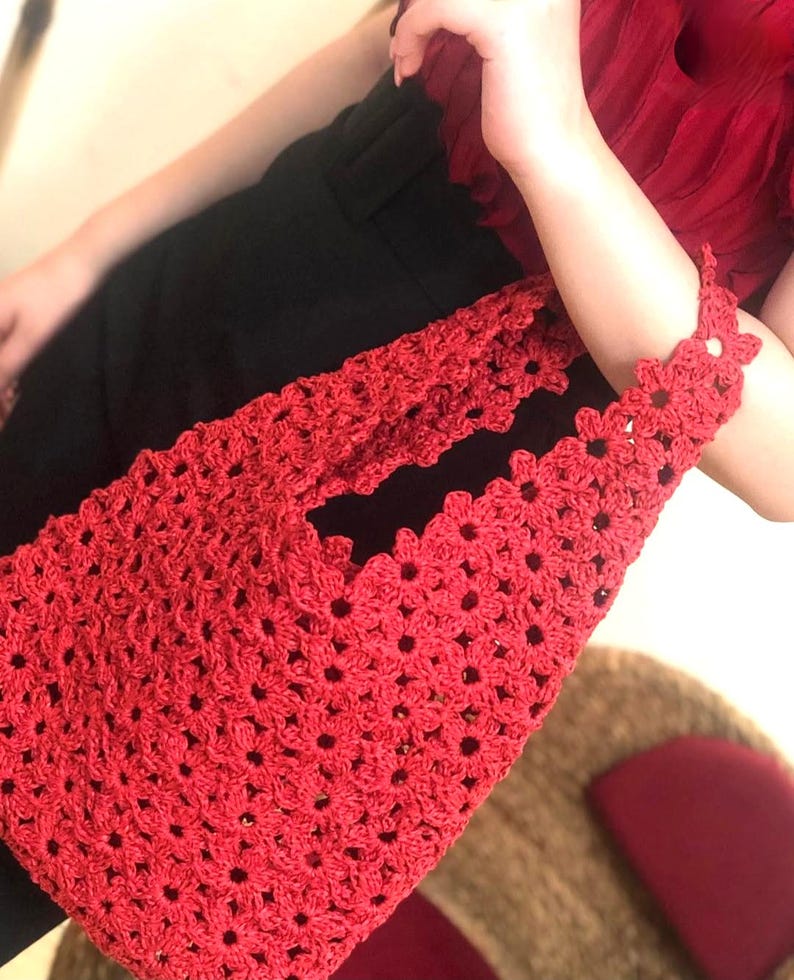 May include: A red crochet tote bag with a floral pattern. The bag is held by a person wearing a red top and black pants. The bag's handle is adorned with flower-shaped crochet details.
