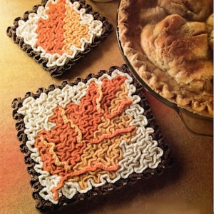 May include: Two crocheted trivets in the shape of a leaf. The trivets are made with brown, orange, and white yarn. The trivets are sitting on a brown surface.