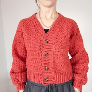 May include: A red crocheted cardigan with a v-neck and button closure. The cardigan has a textured stitch pattern and long sleeves.