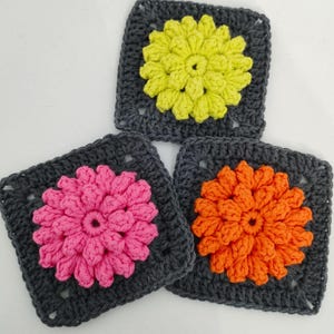 May include: Three crocheted squares with flower designs. Each square has a dark gray border and a central flower in bright colors: yellow, pink, and orange. The flowers have detailed petal patterns, creating a textured appearance.