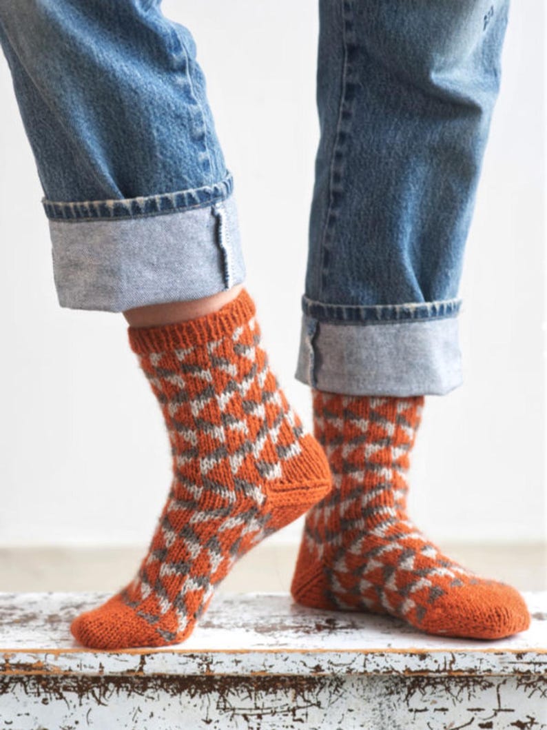 Puede incluir: Un par de calcetines de punto naranja con un patr&oacute;n geom&eacute;trico en blanco y gris. Los calcetines se llevan con vaqueros azules con los pu&ntilde;os doblados, mostrando el dise&ntilde;o de los calcetines. Los calcetines est&aacute;n sobre una superficie blanca.