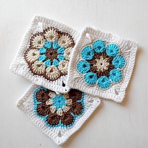 Puede incluir: Tres piezas cuadradas de ganchillo con un diseño floral. Cada cuadrado presenta un borde blanco, con el patrón floral en tonos marrones, beige y azules. El trabajo de ganchillo es detallado y los cuadrados están dispuestos sobre una superficie blanca.