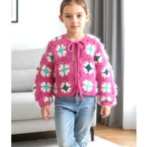 Può includere: Un cardigan rosa all'uncinetto con un motivo floreale, con accenti bianchi, azzurri e blu scuro. Il cardigan ha maniche lunghe e una chiusura con fiocco. La ragazza indossa jeans blu.