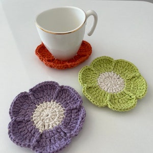 May include: Three crocheted flower-shaped coasters in purple, green, and orange. Each coaster has a white center.