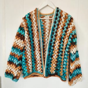 Könnte beinhalten: Handgefertigte Häkeljacke im Granny-Square-Muster, ohne Verschluss. Die Jacke hat eine Farbpalette aus Türkis, Braun, Beige und Creme. Das Kleidungsstück wird auf einem weißen Kleiderbügel vor weißem Hintergrund präsentiert.