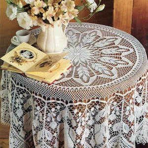 May include: A round, white crocheted tablecloth with a detailed floral pattern covers a wooden table. A white vase with a bouquet of flowers, a teacup, and a small book are on the table. The tablecloth's intricate design is visible.