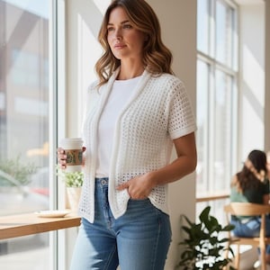 May include: A woman wearing a white, short-sleeved crochet cardigan over a white t-shirt and blue jeans. She is holding a coffee cup. The cardigan has a textured pattern and a draped front. The setting appears to be a cafe or a well-lit room.