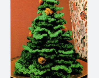 Puede incluir: Un árbol de Navidad tejido a mano, de color verde, con un aspecto texturizado. El árbol está adornado con pequeños adornos redondos amarillos y rojos. Se muestra sobre una superficie redonda de color marrón.