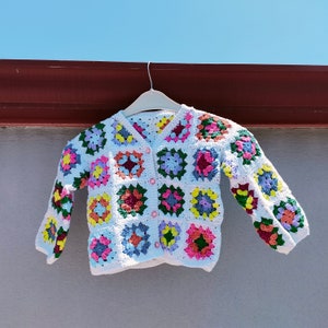 May include: A white crochet cardigan with colourful square patterns. The cardigan has a V-neck, long sleeves, and pink buttons. The squares are in various colours, including pink, yellow, blue, and green, creating a patchwork design.
