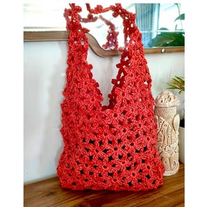 May include: A vibrant red crochet tote bag, featuring a floral pattern. The bag has a handle made of the same crochet material and design. The bag is displayed on a wooden surface.