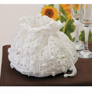 Puede incluir: Un bolso blanco de ganchillo con cordón, de forma redonda. El bolso está adornado con pequeñas perlas de imitación y tiene una textura delicada y calada. El bolso está sobre una superficie marrón oscuro.