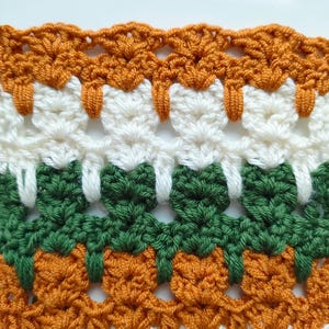 May include: Close-up of a crocheted fabric with alternating rows of orange, white, and green. The fabric has a textured, lacy pattern.